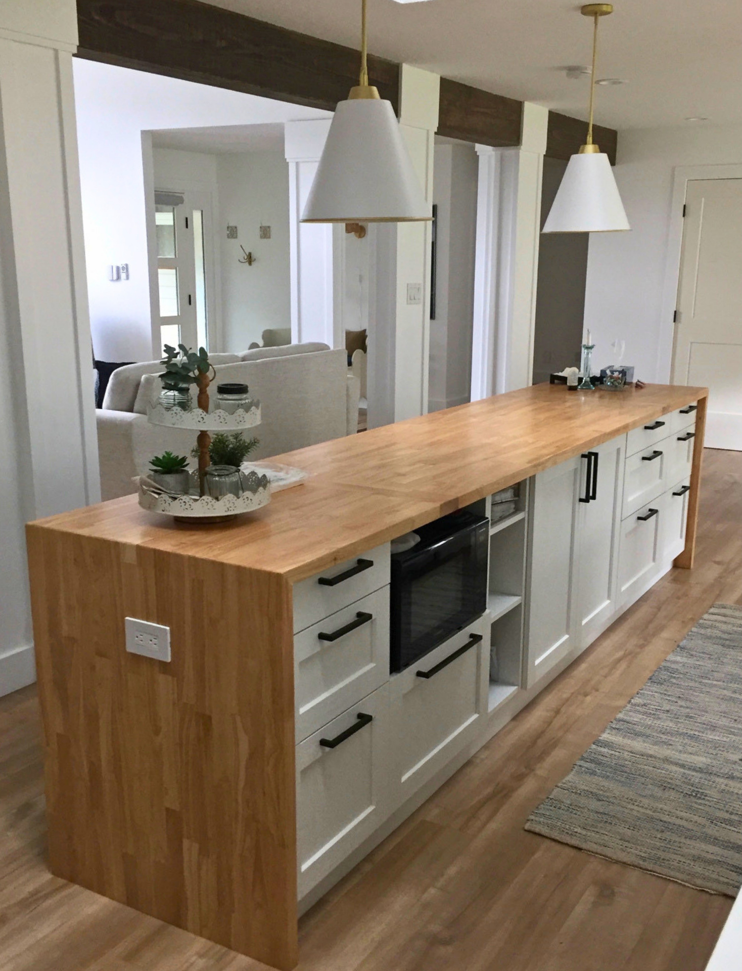 Long white shaker island with butcher-block top, matte-black bar pulls, brass-and-white cone pendants and a dark wood ceiling beam, opening to an adjoining living room — a barrier-free single-level layout suited to aging in place.